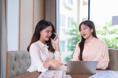 İki genç Asyalı kadın, modern bir ofiste dizüstü bilgisayar ve akıllı telefon kullanarak iş stratejisi analizi yapıyor. Pazarlama eğilimleri ve grafikleri belgesi tartışılıyor.