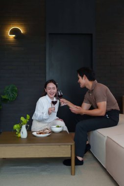 Asyalı bir çift evde kırmızı şarapla kadeh kaldırarak romantik bir akşam geçirir. Yetişkin adam ve genç kadın gülümsüyor, modern odadaki yemek masasında mutlu bir kutlama için..