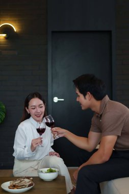 Asyalı yetişkin bir çift kırmızı şarap kadehleriyle kutlama yapıyor. Evde sıcak bir atmosferde akşam yemeği yiyip romantik bir şekilde kadeh kaldırmayı seviyorlar..