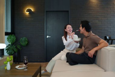 Mutlu Asyalı çift evlerinde rahat vakit geçiriyorlar. Kadın, partnerinin salatasını modern bir oturma odasında besliyor. Sağlıklı yemek ve kaliteli zamanı aşk ilişkileriyle paylaşıyor.