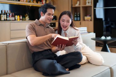 Bu resim, evlerindeki kanepede kitap okuyan yetişkin bir Asyalı çifti gösteriyor. Gülümsüyorlar ve çok mutlu görünüyorlar. Birlikte boş zamanlarının tadını çıkarıyorlar. Bu bir birliktelik ve paylaşılan bir yaşam tarzını ifade eder..