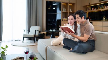 Yetişkin bir Asyalı çift modern oturma odasındaki kanepede kitap okuyarak birlikte kaliteli zaman geçiriyorlar. Evlerinde huzurlu bir atmosfer ve şarapla rahatlatıcı bir akşamın tadını çıkarıyorlar..