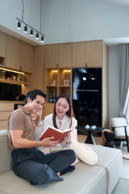 Asyalı bir erkek ve bir kadın, yetişkin bir çift, evde bir kanepede oturmuş birlikte kitap okuyorlar. Rahatlıyorlar, gülümsüyorlar ve modern evlerinde boş zamanlarının tadını çıkarıyorlar..