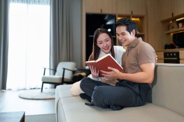 Genç Asyalı çift kanepede birlikte kitap okuyor. Onlar modern evlerinde gülümsüyor ve kaliteli zaman geçiriyorlar. Bu, rahatlatıcı sevgi dolu bir ilişkiyi ve konforlu bir yaşam tarzıyla aile bağını tasvir eder..