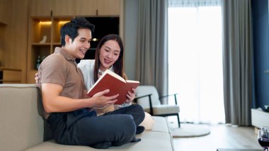 Genç bir Asyalı çift rahat oturma odalarında rahat bir kanepede birlikte kitap okuyorlar. Ev hayatı konseptlerinde rahat ve samimi bir anın tadını çıkarıyorlar..