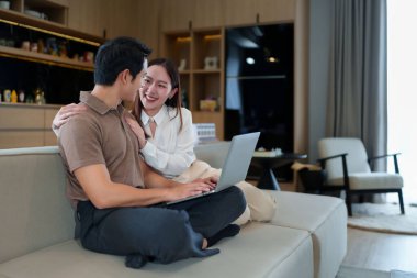 Asyalı genç çift evde kanepede dizüstü bilgisayar kullanıyor. Gülümsüyor ve eğleniyorlar. Bu resim modern yaşam tarzını ve konfor ve bağlantıyla olan ilişkileri gösterir..