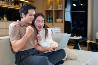 Yetişkin bir Asyalı çift evdeki kanepede dizüstü bilgisayar kullanırken heyecanlanır. Birlikte tezahürat yapıp başarılarını kutlarlar. Bu mutlu an neşe ve heyecanla doludur.