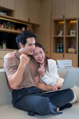 Evde kazandığımız zaferi kutlamak için dizüstü bilgisayar kullanan heyecanlı bir Asyalı çift. Gülümsüyorlar, neşeyle ekrana bakıyorlar, online çalışmanın güzel bir başarısı.