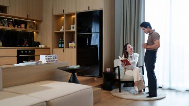 Yetişkin bir Asyalı çift modern dairelerinde dinlenmenin tadını çıkarıyor. Kadın oturmuş kitap okuyor ve adam açık bir alanda kahve içiyor..