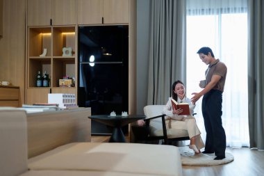 Bu görüntü, modern evlerinde yaşayan Asyalı bir çiftin iç açıcı bir sahnesini çekiyor. Yetişkin kadın, elinde kahve fincanıyla kitap okuyor. Sıcak, sevgi dolu bir ortam yaratıyorlar.