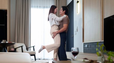 Genç bir Asyalı çift modern oturma odalarında sevgi dolu bir şekilde kucaklaşıyor, birlikte tatlı ve hassas bir anın tadını çıkarıyorlar, sıcak ve sevgi dolu bir ortam yaratıyorlar.