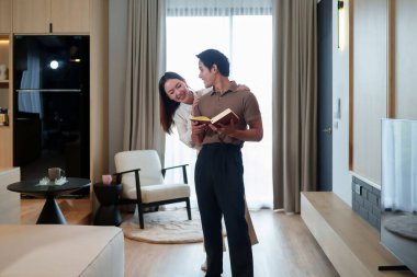 Sevgi dolu bir Asyalı çift, evinde özel bir anı paylaşıyor. Kadın, erkeğin parlak oturma odasında kitap okumasını kucaklıyor. Rahat, modern oturma odasını doğal ışıkla yakalıyor..