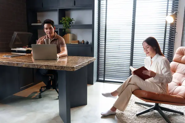 Evde çalışan iki yetişkin Asyalı adam, mermer bir masada dizüstü bilgisayar, sandalyede kitap okuyan bir kadın. Konforlu ev ofisi ve çalışma ortamı.