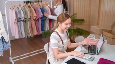 Yetişkin Asyalı kadın bilgisayarını ve hesap makinesini kullanarak online kıyafet mağazasını yönetiyor. Siparişlerle, finansmanla ilgileniyor ve e-ticaret işinin titiz bir planlamayla gelişmesini sağlıyor..