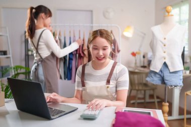 Önlüklü Asyalı genç kadınlar, hesap makinesi ve satış ödemeleri için dizüstü bilgisayarlarıyla çevrimiçi giyim mağazasını yönetiyorlar. Stüdyoda çalışan küçük bir girişimci..