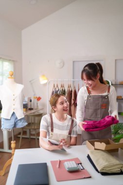 İki Asyalı kadın gülümsüyor ve çevrimiçi giyim mağazalarında birlikte çalışıyorlar. Müşteri siparişleri paketliyorlar. Ev sahibi girişimcilikten zevk alıyor.