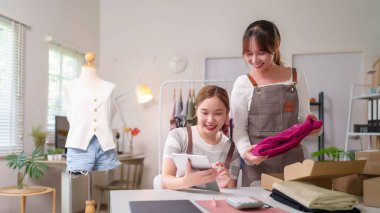 İki gülümseyen Asyalı genç kadın küçük bir online moda şirketine sahipler. Tablet siparişlerini kontrol ediyorlar ve giysileri kutulara dolduruyorlar. Müşterilere gönderilmeye hazır. Başlangıç konsepti.