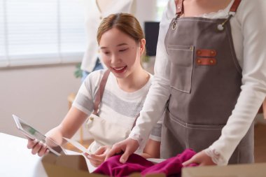 İki Asyalı genç kadın, stoklarını kontrol etmek için bir tablet kullanarak ve müşterilerinin siparişlerini dikkatle ve dikkatle nakliye için hazırlayarak çevrimiçi giyim mağazalarını yönetiyor..