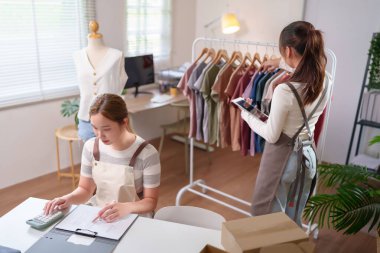 Bu görüntü Asyalı kadın girişimcilerin online moda mağazalarını yönettiklerini gösteriyor. Stok yapıyorlar, siparişleri hesap makinesi ve tabletle kontrol ediyorlar. Evden çalışmak küçük bir iştir..