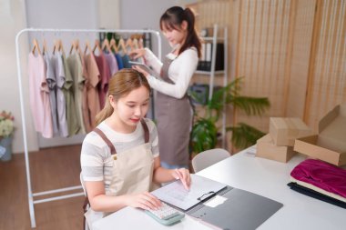 İki genç Asyalı kadın online bir giyim mağazası işletiyor, envanter yönetimi için bir hesap makinesi kullanıyorlar ve perakende e-ticaret başarısı siparişlerini kontrol ediyorlar..