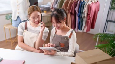 İki gülümseyen Asyalı kadın online moda işi yapıyor. Envanter sayımı için bir tablet kullanıyorlar ve ecommerce girişimi için kutu paketleyerek telefon siparişi alıyorlar.