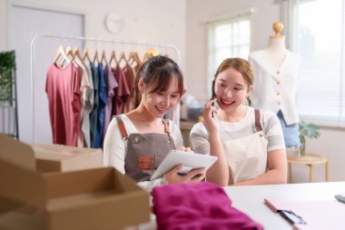 İki genç Asyalı kadın online moda mağazalarını yönetiyor. Biri siparişleri kontrol etmek için tablet kullanırken, diğeri siparişleri doğrulamak için bir müşteriyle cep telefonuyla konuşuyor..