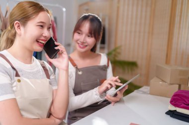 İki Asyalı genç kadın online işlerinde birlikte çalışıyorlar. Biri telefonda, diğeri de tabletten siparişleri kontrol ediyor. Gülümsüyor ve girişimcilikten memnun..