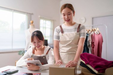 İki genç Asyalı kadın birlikte çalışıyor, online moda işlerini yönetiyorlar. Siparişleri paketliyorlar ve stokları kontrol etmek için bir tablet kullanıyorlar. Müşterilere dikkatli bir şekilde teslim edilmesini sağlıyorlar..