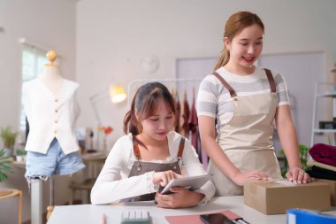 İki genç Asyalı kadın çevrimiçi siparişleri kontrol ediyor ve küçük işletmelerinde sevkiyat için paketler hazırlıyorlar. Online pazarlama için paket kutuları ve diğer malzemelerle çalışıyorlar.