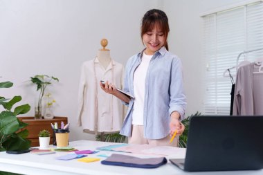 Genç Asyalı kadın yeni kıyafet tasarımları üzerinde çalışıyor. Etrafı kumaş örnekleri ve diğer tasarım materyalleriyle çevrili. Moda tasarım şablonu oluşturma ve dikme işlemi