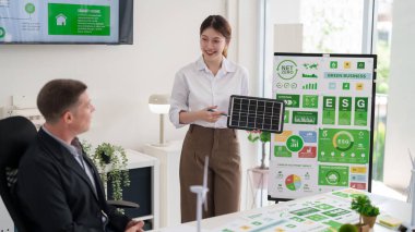 Asya 'lı genç bir kadın ESG toplantısında beyaz bir erkeğe güneş paneli sunarak net sıfır karbon emisyonu ve akıllı ev çözümlerini tartışıyor.