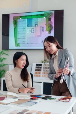İki genç Asyalı kadın iç mimar, parlak bir ofis alanında tasarım fikirlerini tartışan planlar ve renk paletlerini gözden geçiren bir ev yenileme projesinde işbirliği yapıyorlar.