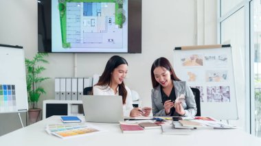 İki genç Asyalı kadın iç mimar birlikte çalışıyor, renk paletleri seçiyorlar ve modern ofis ortamında dizüstü bilgisayar kullanarak ev yenileme çalışmaları planlıyorlar..