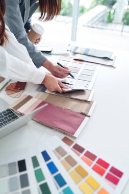 İki Asyalı kadın bir iç tasarım projesi için kumaş ve renk paletlerini gözden geçiriyorlar. Eller beyaz masadaki kumaş örneklerini seçmek için kalem kullanıyor..