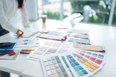 Asyalı yetişkin bir kadın iç mimar renk şeması seçiyor. Renk örnekleri, tekstil örnekleri ve tasarım planları parlak beyaz ofis masasında düzenlenir..