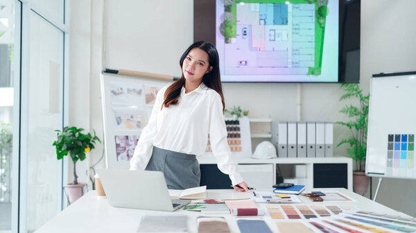 Young Asian woman architect working on a design project in a modern office space with blueprint and color swatches. She is working on interior project.