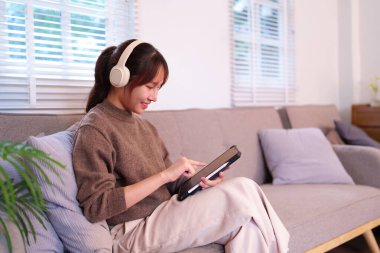 Genç bir Asyalı kadın yapay zeka tableti ile etkileşime girerken son derece odaklanmış ve gülümsüyor. Oturma odasındaki kanepede kulaklık takıyor. Bu sahne modern günlük yaşam, eğlence ve teknolojiyi resmediyor..