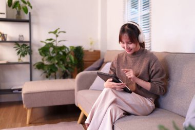Bu görüntü, tabletiyle rahatça meşgul olan Asyalı genç bir kadını yakalıyor. Kablosuz kulaklık takıyor. İlginç bir deneyim için. Ev düzeni teknolojiyle geliştirilmiş modern yaşamı yansıtıyor. Bu sahne, günlük yaşamı smayla betimler.
