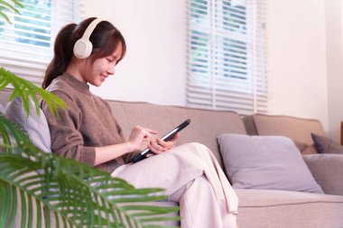 Rahat genç Asyalı kadın kulaklık takarken tablet üzerinde dijital içerikle meşgul oluyor. Rahat bir ev ortamında modern günlük yaşam teknolojisinin tadını çıkarıyor.