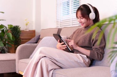 Genç bir Asyalı kadın bir kanepede dinleniyor ve tabletine bir stil kullanarak bağlanıyor. Beyaz kulaklıklar ilgi çekici sesler sağlıyor. Bu samimi sahne, kişisel teknolojiyi ve günlük yaşamına kusursuz yapay zeka entegrasyonunu vurguluyor..