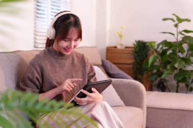 Bu resim, modern teknolojiden zevk alan genç bir Asyalı kadını evdeki dijital etkileşim için kulaklıklarıyla tabletini kullanırken gösteriyor.
