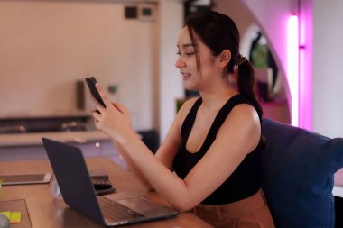 Genç Asyalı kadın dijital göçebe canlı yaratıcı ofis ortamında telefon ve taşınabilir bilgisayarla uğraşırken mutlu bir şekilde gülümsüyor.