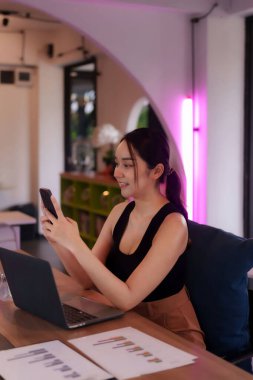 Yetişkin Asyalı kadın şık bir çalışma alanında akıllı telefon ve dizüstü bilgisayar kullanıyor. Dijital görevlere ve uzak çalışma verimliliğine odaklanmış.