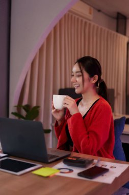 Neşeli Asyalı kadın serbest çalışan modern ortak bir ofiste dizüstü bilgisayara odaklandı. Kahve içiyor, üretken ve esnek hissediyor..