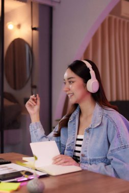 Yetişkin bir Asyalı kadın modern bir çalışma alanında verimli çalışıyor. Bir proje için notlar alırken kulaklıkla dinliyor..