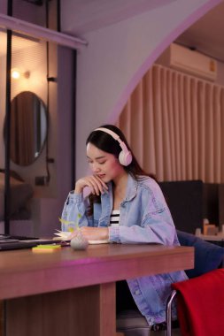 Asyalı kadın, modern bir çalışma alanında özenle çalışıyor. Derinden odaklanmış görünüyor, not alırken kulaklık takıyor. İdeal uzak çalışma ayarları.