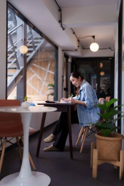 Asyalı yetişkin kadın çalışkan bir çalışma alanında özenle çalışıyor. O, kitabında yaratıcılığı ve verimliliği teşvik eden notlar yazmaya odaklanıyor..