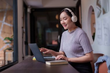 Yetişkin Asyalı kadın, dinamik bir ortamda iş amaçlarına ulaşmak için dizüstü bilgisayar ve kulaklık kullanarak modern bir çalışma alanında mutlu bir şekilde çalışıyor.
