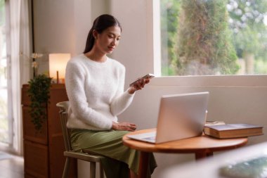 Asyalı genç bir kadın akıllı telefonu ve laptopuyla evde güneşli bir pencerenin yanında huzurlu bir ortamın tadını çıkarıyor.