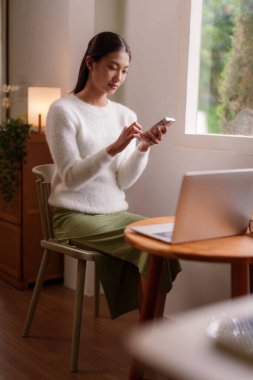 Asyalı yetişkin bir kadın sıcak oturma odasında akıllı telefonuyla barışçıl bir şekilde iletişim kuruyor. Rahat bir atmosferde çevrimiçi bağlanıyor..